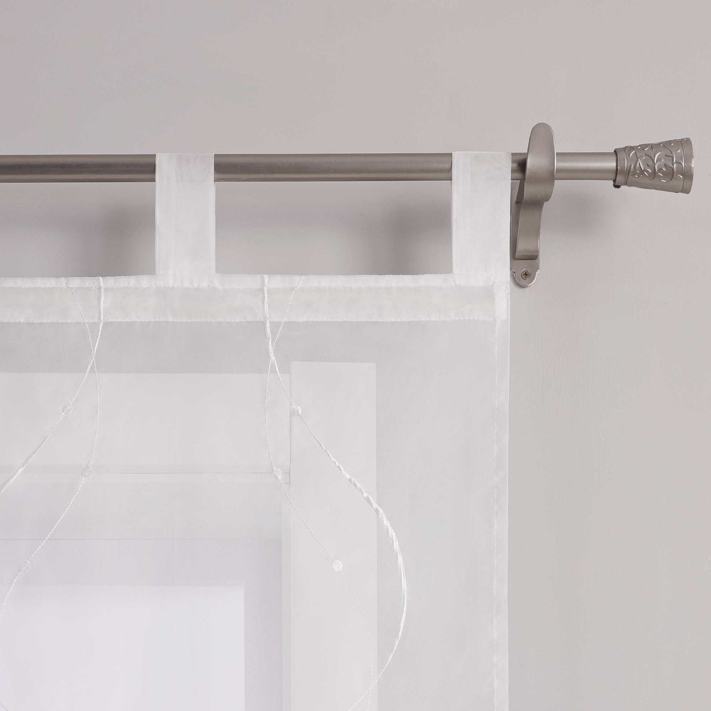Das Bild zeigt die Details der Schlaufen einer weissen Gardine. Die Gardine ist mit feiner weisser Naht in Wellenform und kleinen Punkten am Ende verziert. Sie haengt mithilfe gleichmaessiger weisser Schlaufen an einer Metallstange. Die Naehte sind sauber verarbeitet und das Material wirkt hochwertig.