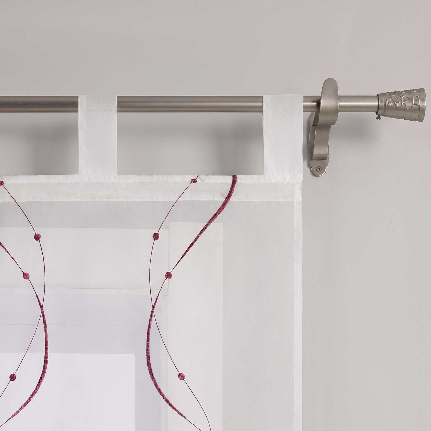 Auf dem Bild ist ein Detail der Schlaufen einer weissen Gardine zu sehen. Die Gardine ist mit feinen, lila wellenfoermigen Naehten versehen, deren Enden kleine Punkte bilden. Die Gardine ist mithilfe gleichmaessiger weisser Schlaufen an einer Metallstange aufgehaengt. Die Naehte sind sauber verarbeitet, das Material wirkt hochwertig.