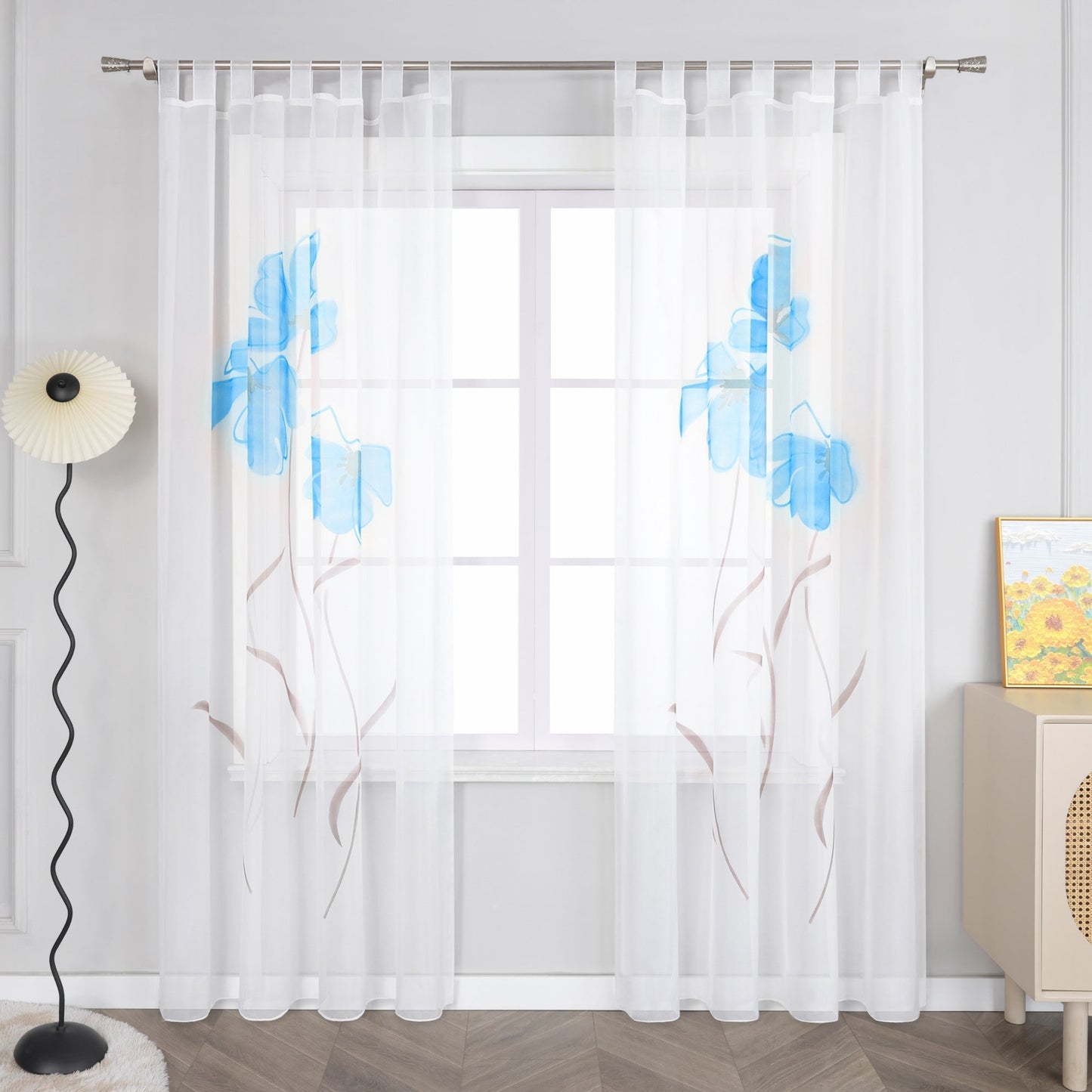 Das Bild zeigt einen Vorhang aus dünnem, weißen Stoff mit blauen Blumenmustern. Der Vorhang hat Riegel an der Oberseite, um ihn an einer Gardinenstange aufzuhängen. Neben dem Vorhang steht eine einzigartige Stehlampe, und auf dem kleinen Schrank rechts hängt ein Gemälde in Gelbtönen.