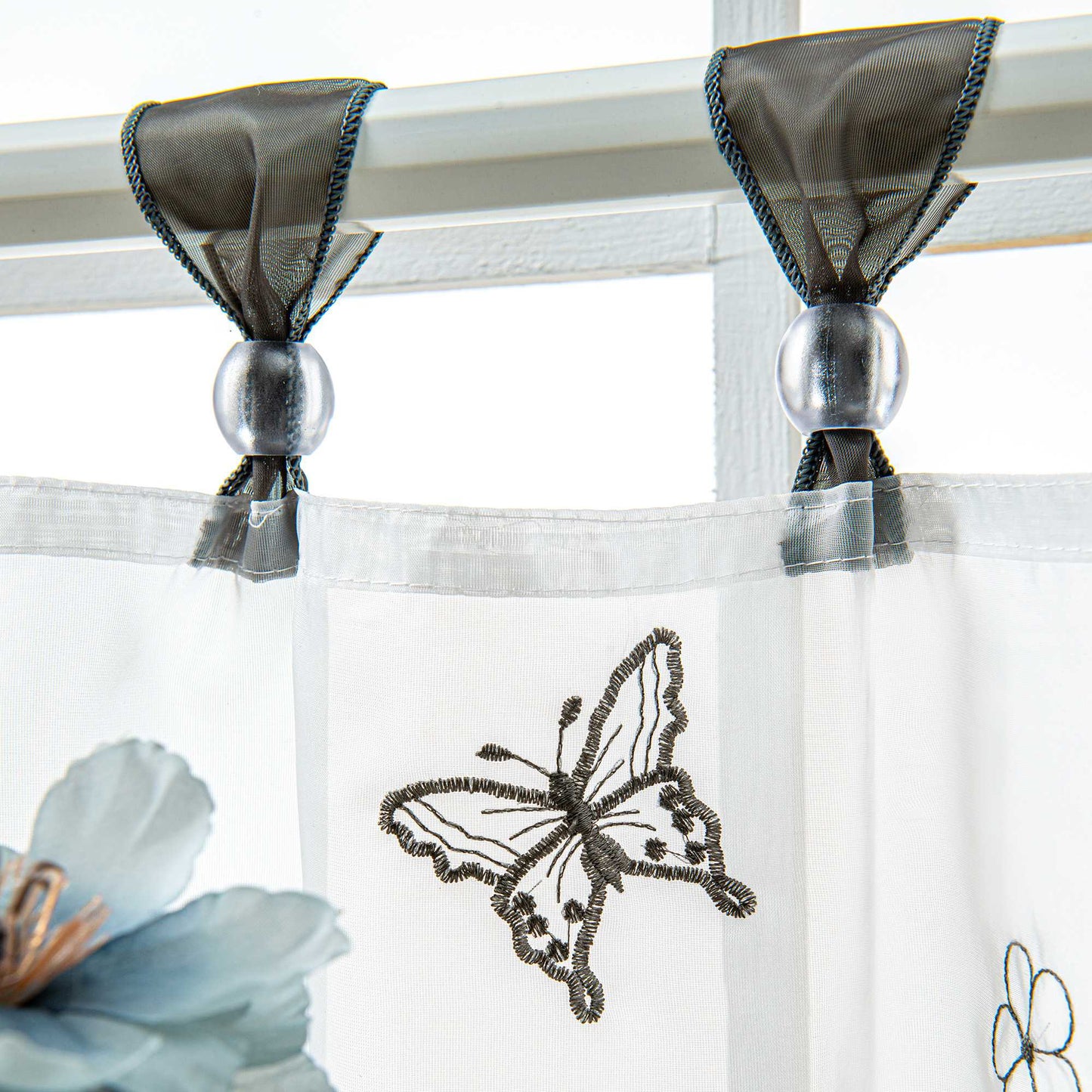 Nahaufnahme der grauen Vorhangringe mit transparenten Perlen, der Vorhang hängt an einer Stange und zeigt Schmetterlingsmuster, daneben sind Blumen als Dekoration.