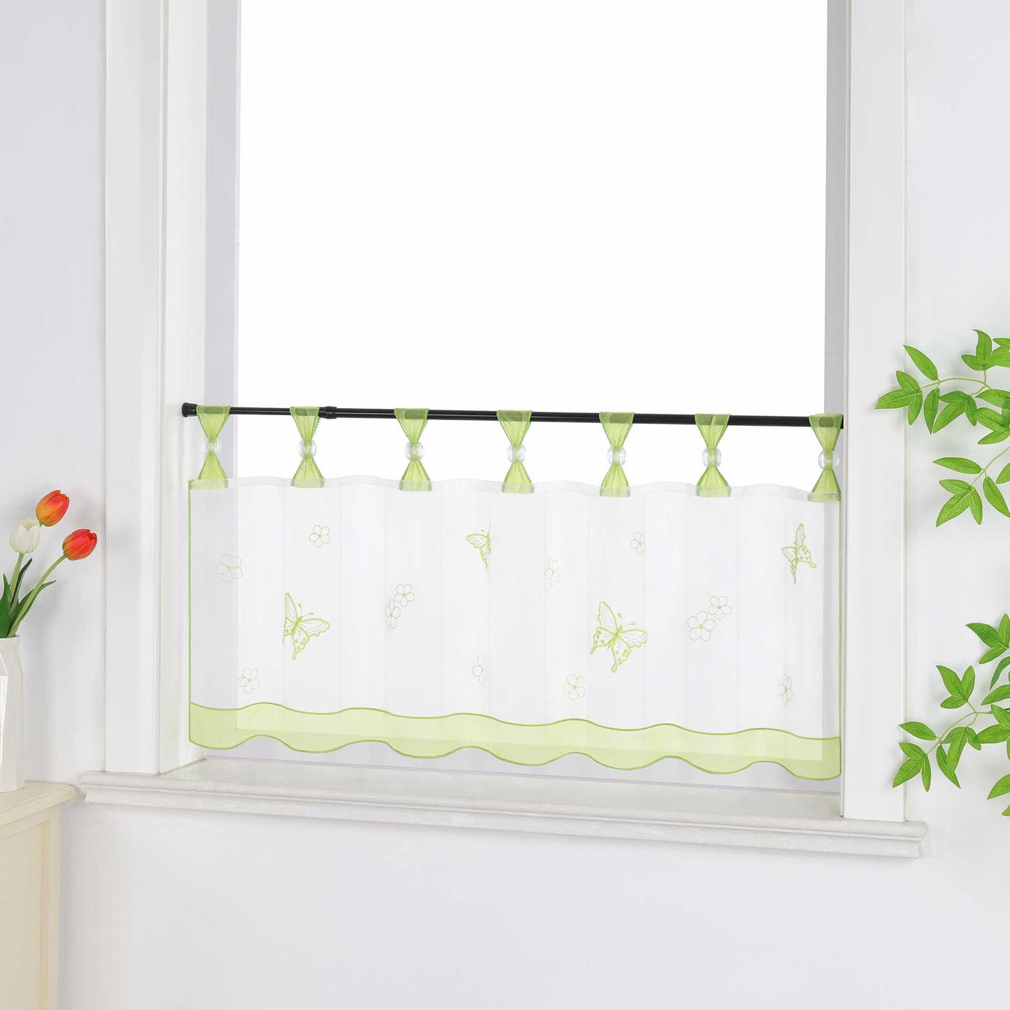 Seitliche Ansicht eines gruenen transparenten Kurzgardine mit dekorativen Perlen an den Aufhängeschlaufen und Schmetterlingsmuster auf der Oberfläche. Der Vorhang hat ein gruenes Dekoband am unteren Rand und ist vor einem Fenster mit einem Schrank und Blumen als Dekoration aufgehängt.