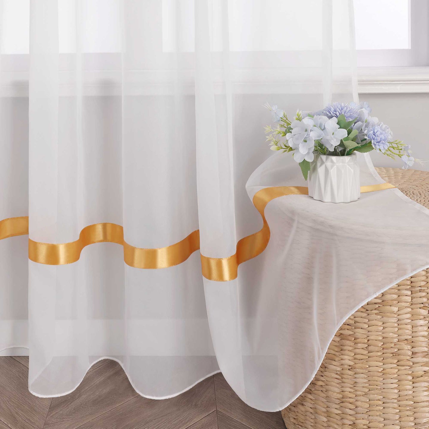 Am Fenster haengt ein weisser, halbtransparenter Vorhang mit orangefarbenen Satinbaendern aus Voile. Darauf steht eine weisse Vase mit einem Strauss aus blau-weissen Blumen.