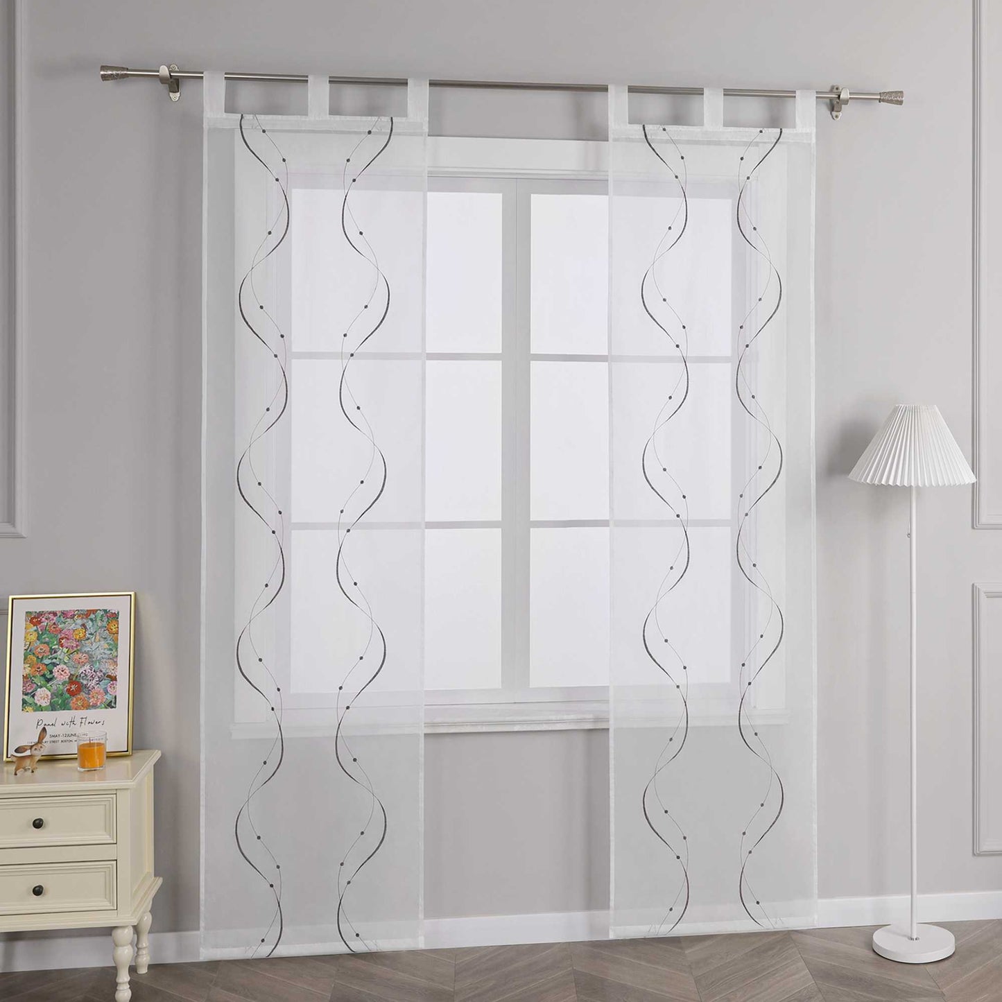 Joyswahl – Das Bild zeigt die Seitenansicht einer weissen transparenten Gardine mit Wellenperlenmuster und Stickerei, die an einer Metallstange haengt. Links vom Fenster steht ein heller Schubladenschrank mit einem farbigen Blumenbild und einem kleinen orangen Objekt darauf. Rechts vom Fenster steht eine Stehlampe mit einem weissen gestreiften Lampenschirm.