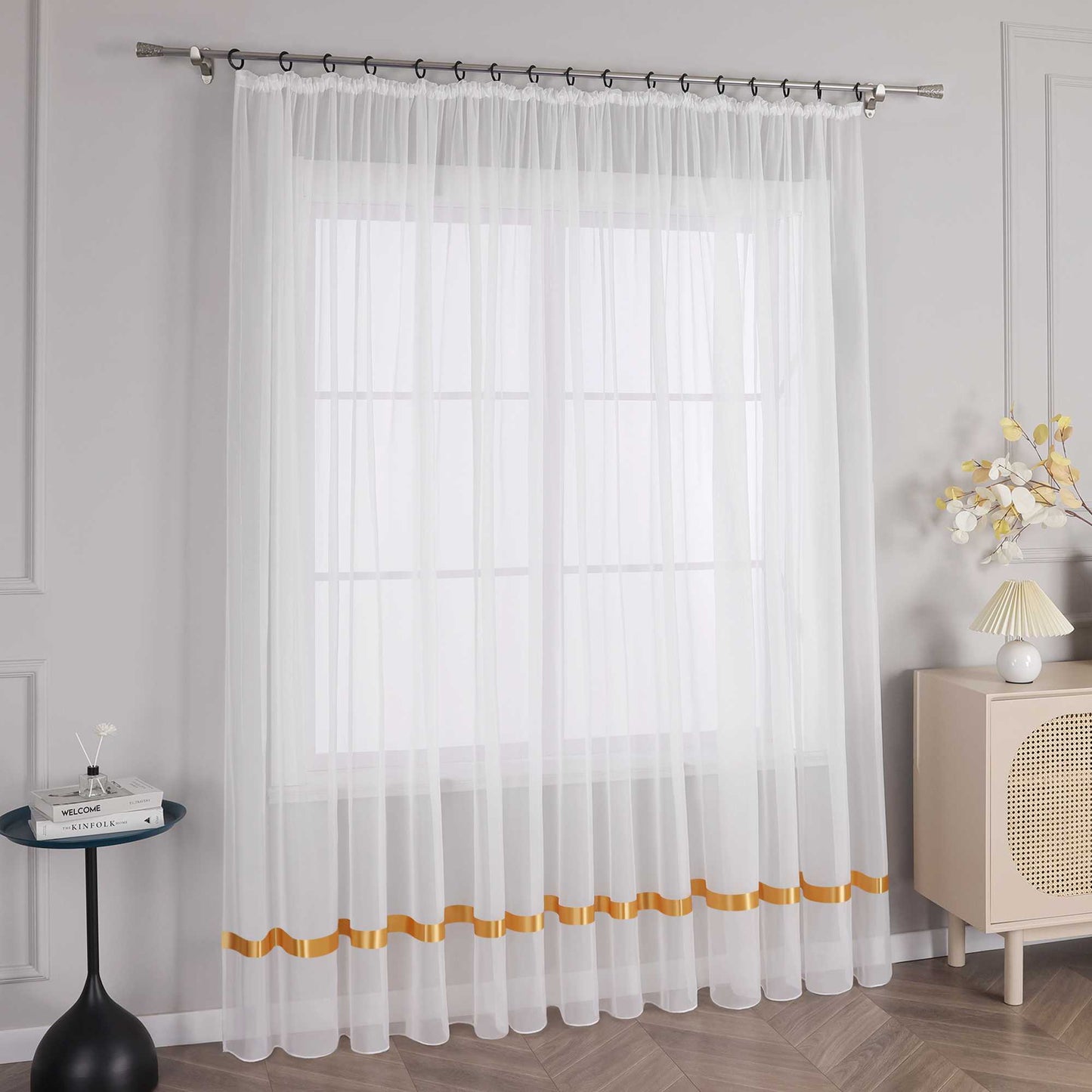 An einem Fenster haengt ein weisser Vorhang mit orangefarbenen Satinbaendern und Kraeuseln. Am oberen Ende befindet sich eine Metallstange mit schwarzen Ringen, die den Vorhang an seinem Platz halten. Auf der linken Seite des Fensters steht ein kleiner schwarzer runder Tisch mit einigen Gegenstaenden. Rechts vom Fenster steht ein heller Schrank mit einer Lampe mit hellem Schirm und einem Strauss dekorativer Blumen.