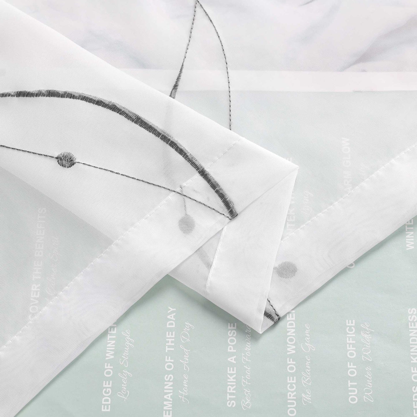 Ein weisser, duenn gewebter Stoff mit schmalem Wellenlinienmuster ist mehrfach gefaltet. Im Hintergrund befindet sich Papier mit aufgedrucktem Text und Motiven.
