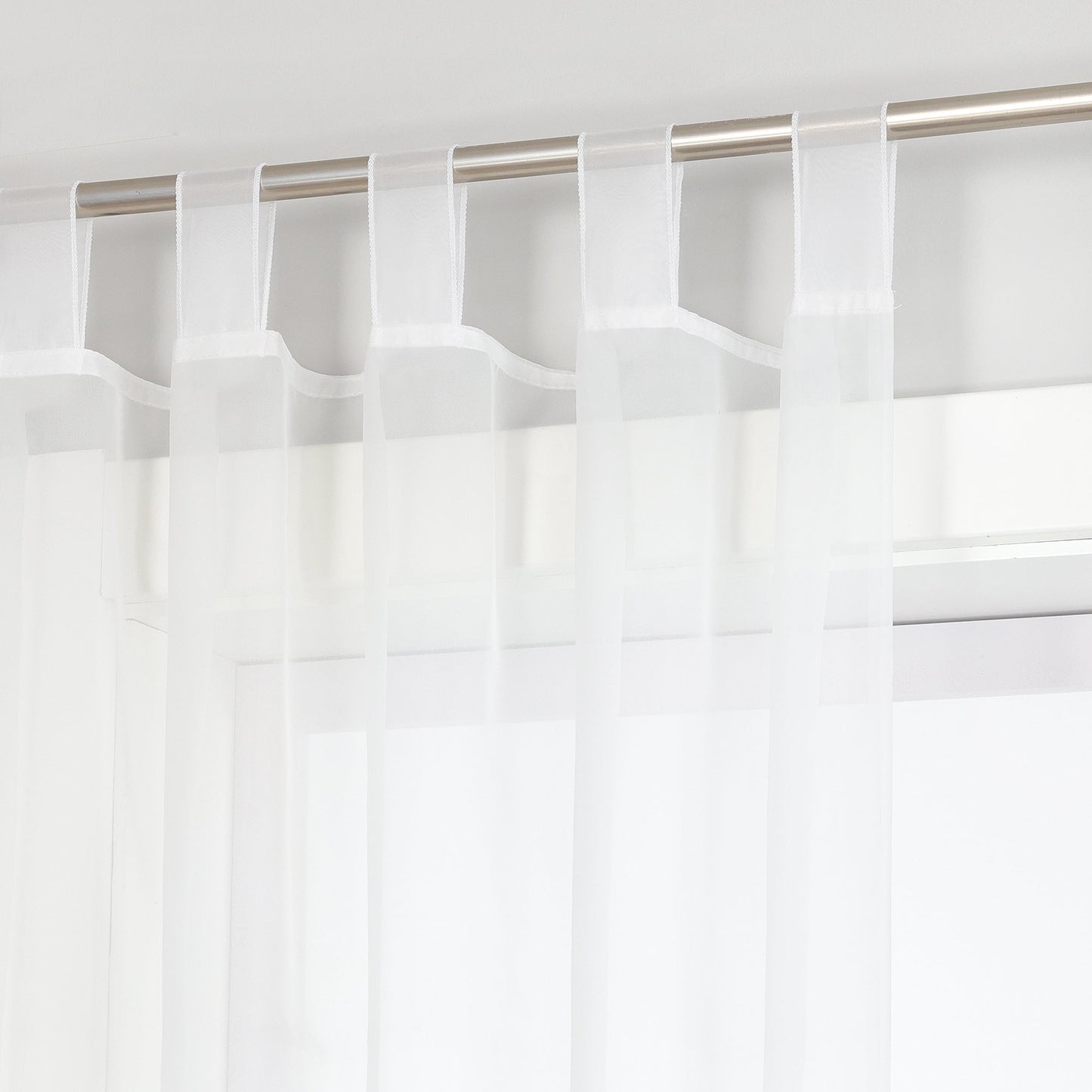 Das Bild zeigt eine Vorhangstange, an der weiße Voile-Vorhänge hängen. Die Vorhänge sind mit Haken an der Stange befestigt, und durch die Voile-Vorhänge ist ein Teil des Fensters sichtbar. Insgesamt vermittelt das Bild einen einfachen und hellen Eindruck.