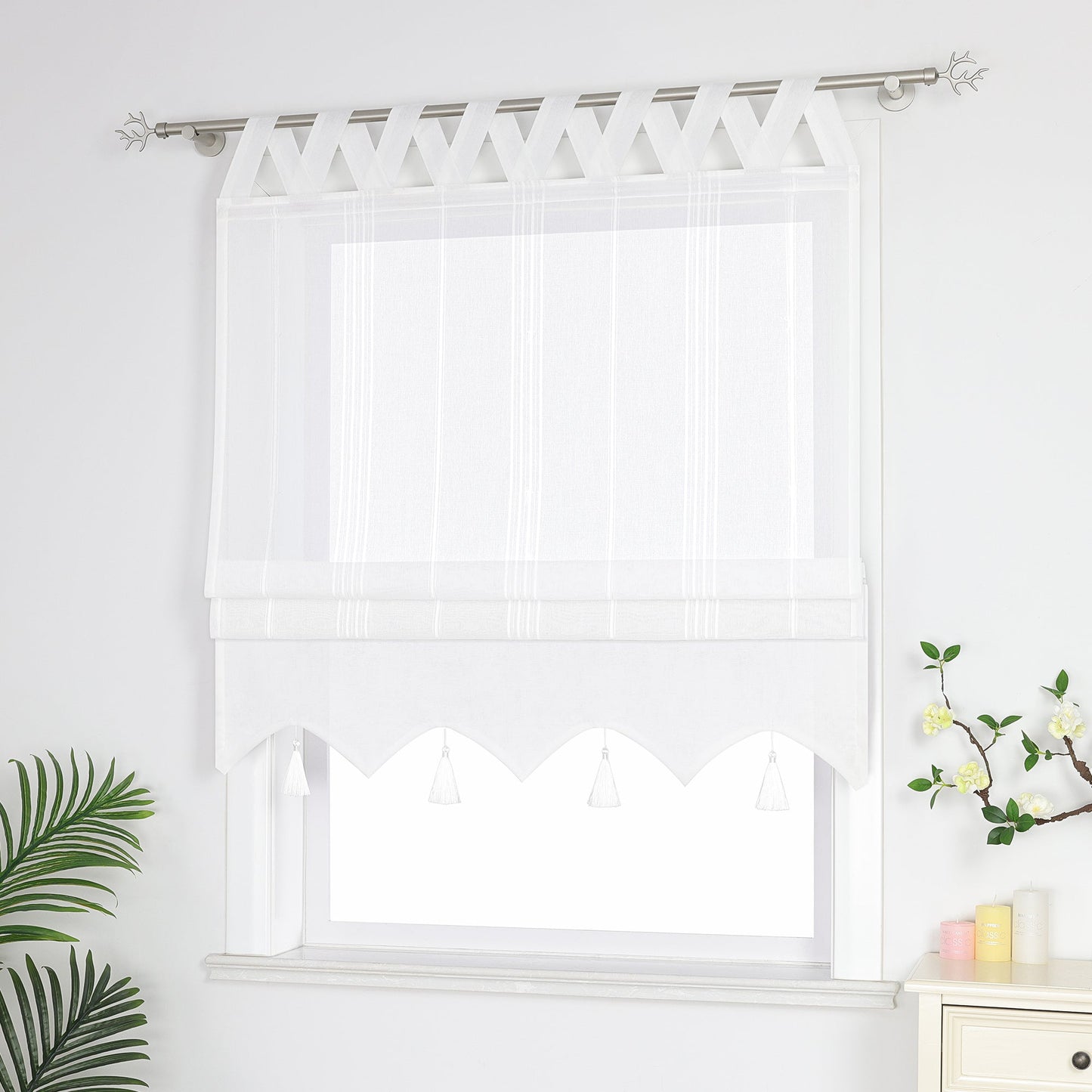 Seitenansicht eines weißen Raffrollos mit Quasten und Schlaufen – halb geschlossen, sodass die Fenster nicht vollständig bedeckt sind, neben Grünpflanzen und duftenden Ornamenten.