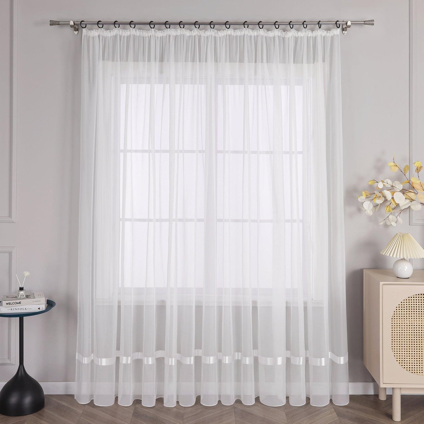An einem Fenster hängt ein Vorhang mit Kräuseln, und am oberen Ende des Vorhangs befindet sich eine Metallstange mit schwarzen Ringen, die den Vorhang an seinem Platz halten. Auf der linken Seite des Fensters steht ein kleiner schwarzer runder Tisch, auf dem einige Gegenstände stehen. Rechts vom Fenster steht ein heller Schrank mit einer Lampe mit hellem Schirm und einem Strauß dekorativer Blumen.