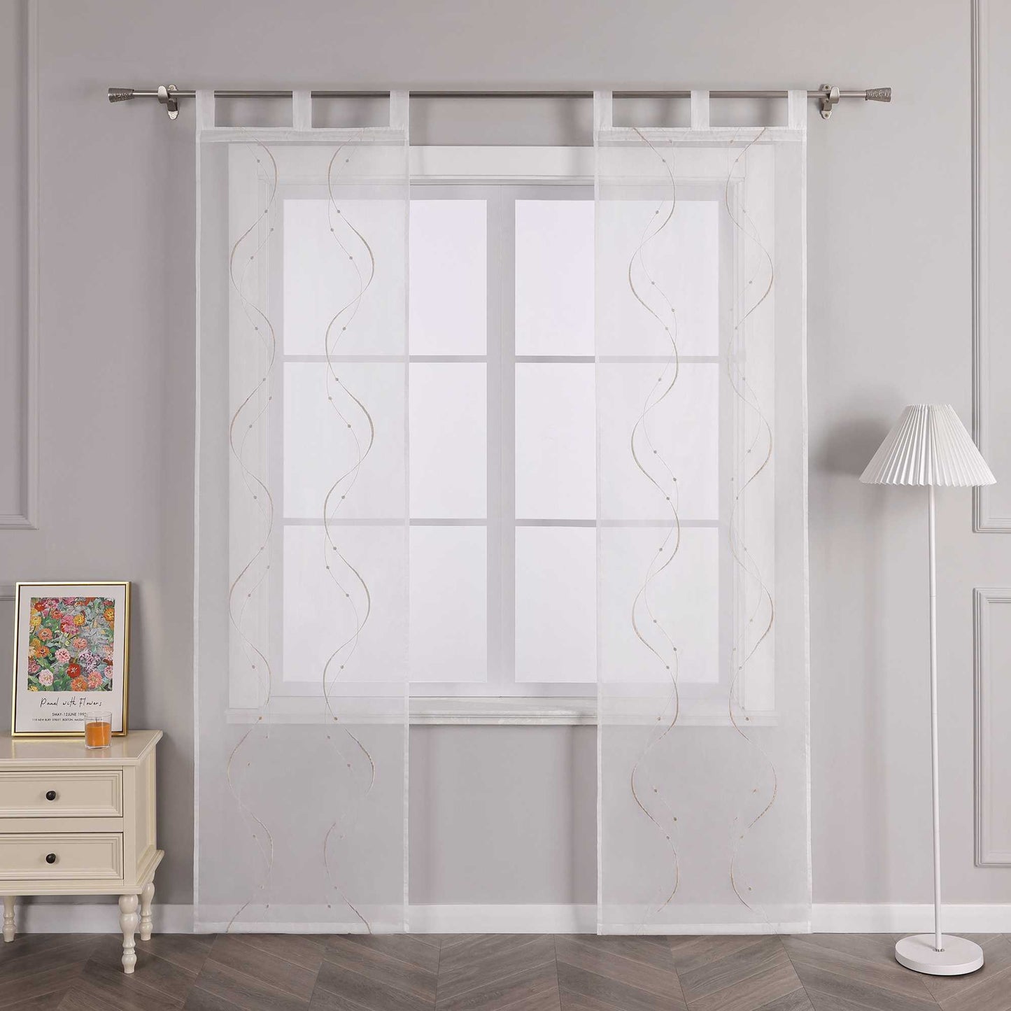 Vor einer hellgrauen Wand befindet sich ein Fenster, an dem eine weisse transparente Schiebe-Gardine mit sandfarbenen symmetrischen Wellenperlenmustern und Stickerei hängt. Die Gardine ist an einer Metallstange mit Schlaufen befestigt. Links vom Fenster steht ein heller Schrank mit Schubladen, auf dem ein farbenfrohes Blumenbild und ein orangenes kleines Objekt stehen. Rechts vom Fenster befindet sich eine Stehlampe mit einem weissen Streifenlampenschirm.