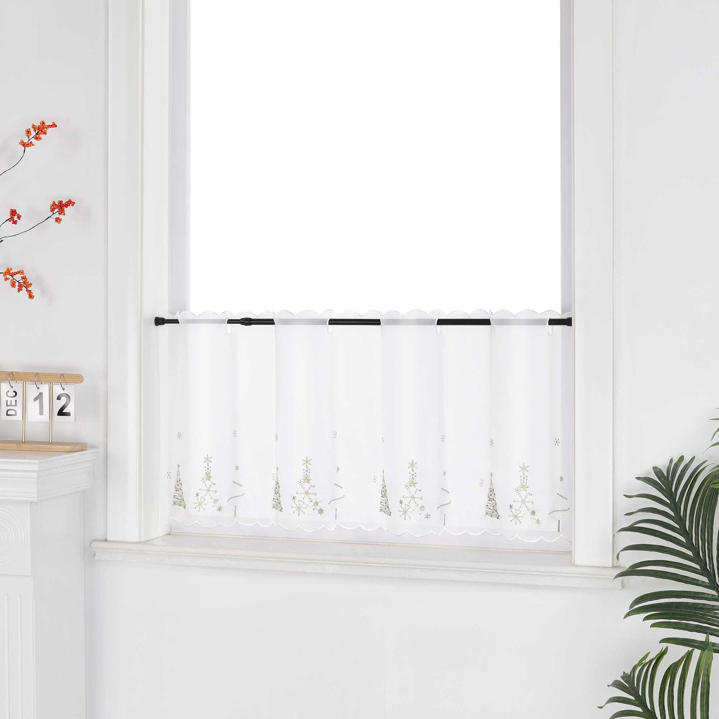 Eine weiße, transparente Scheibengardine mit goldenen Tannenbaum-Motiven hängt vor einem weißen Fenster. Die Wand ist ebenfalls weiß, was den hellen und festlichen Charakter unterstreicht. Seitlich sind dekorative Blumenarrangements zu sehen, die dem Bild eine natürliche und stimmungsvolle Note verleihen – ideal als stimmige Weihnachtsdekoration.