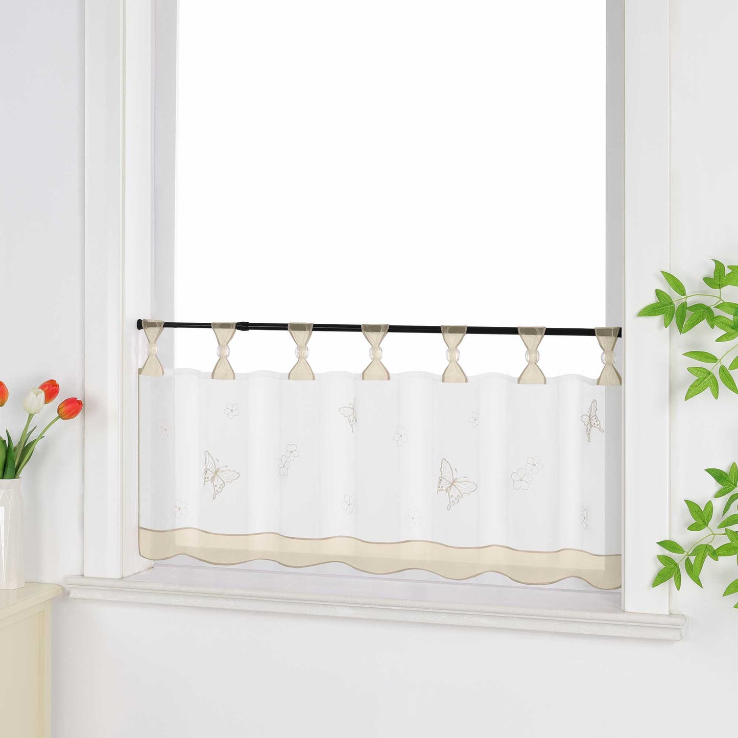 Seitliche Ansicht eines beige transparenten Kurzvorhangs mit dekorativen Perlen an den Aufhängeschlaufen und Schmetterlingsmuster auf der Oberfläche. Der Vorhang hat ein beiges Dekoband am unteren Rand und ist vor einem Fenster mit einem Schrank und Blumen als Dekoration aufgehängt.