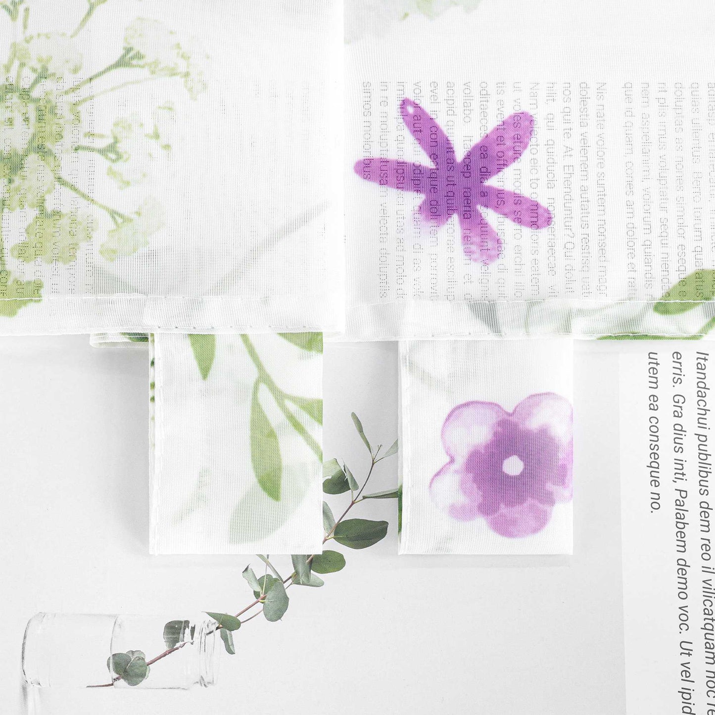Ein Stoff mit Pflanzen- und Blumenmuster, darunter lila Blumen und grüne Zweige. Rechts steht ein transparenter Glasvase mit Eukalyptuszweigen.