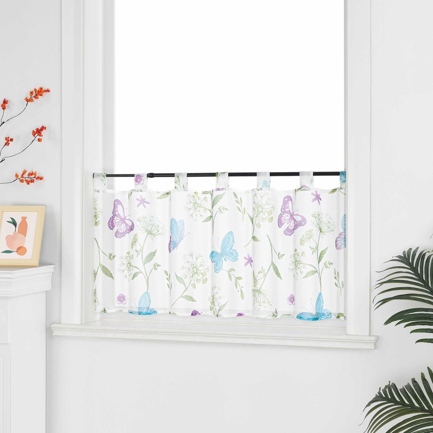 Ein Fenster an einer weißen Wand, mit einer weißen Scheibengardine. Die Gardine zeigt lila und blaue Schmetterlinge, grüne Zweige und weiße Blumen. Auf der linken Seite des Fensters befindet sich ein eingerahmtes Gemälde von Früchten, darüber ein dekorativer Ast mit roten Früchten. Auf der rechten Seite des Fensters stehen grüne Pflanzen.
