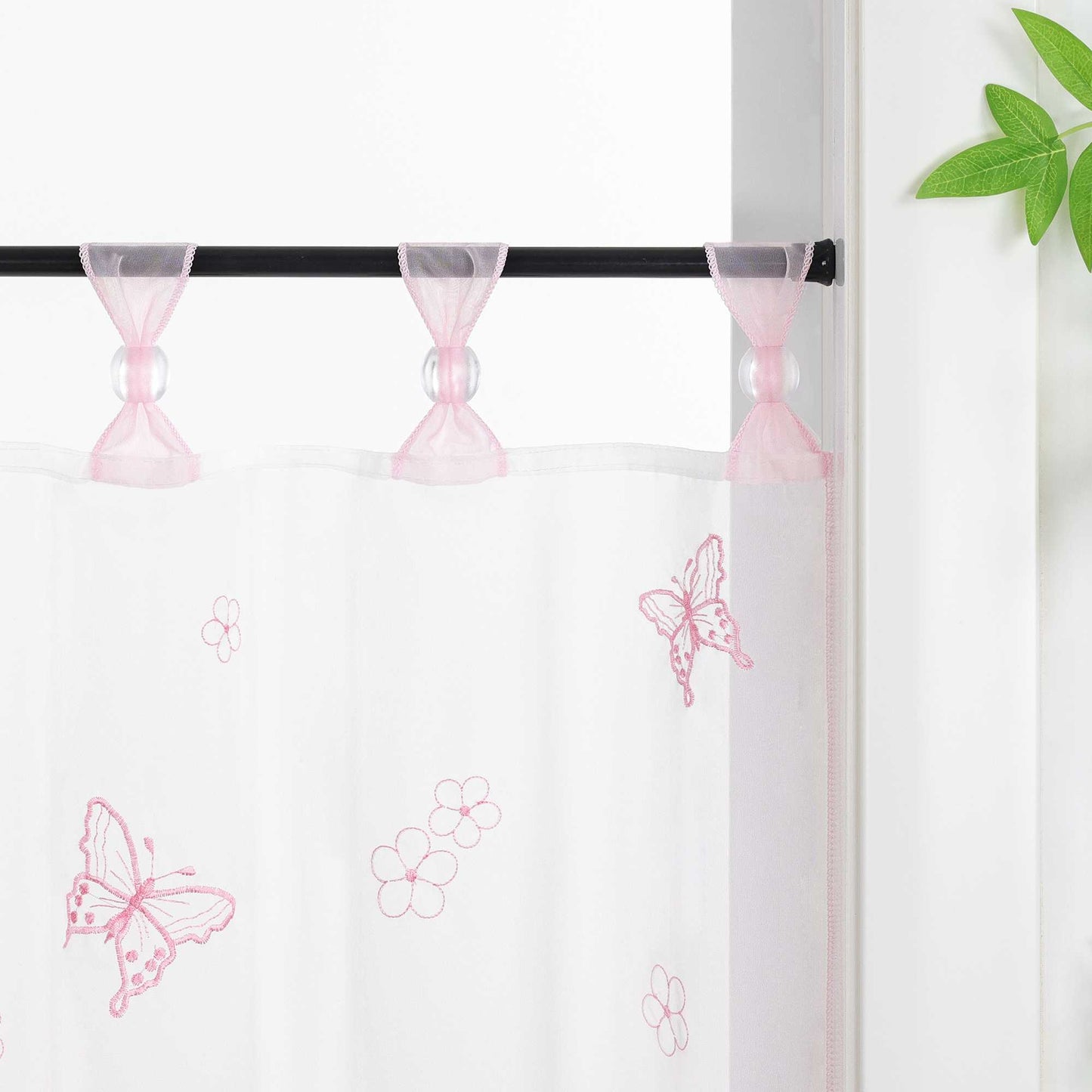 Nahaufnahme der rosa Vorhangringe mit transparenten Perlen, der Vorhang hängt an einer Stange und zeigt Schmetterlingsmuster, daneben sind Blumen als Dekoration.