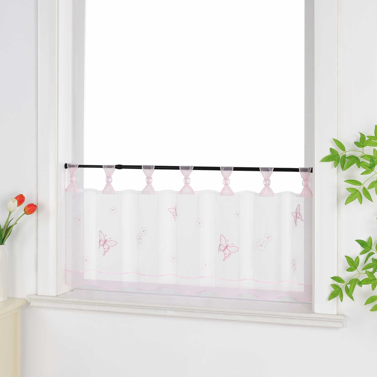 Seitliche Ansicht eines rosa transparenten Kurzvorhang mit dekorativen Perlen an den Aufhängeschlaufen und Schmetterlingsmuster auf der Oberfläche. Der Vorhang hat ein rosa Dekoband am unteren Rand und ist vor einem Fenster mit einem Schrank und Blumen als Dekoration aufgehängt.