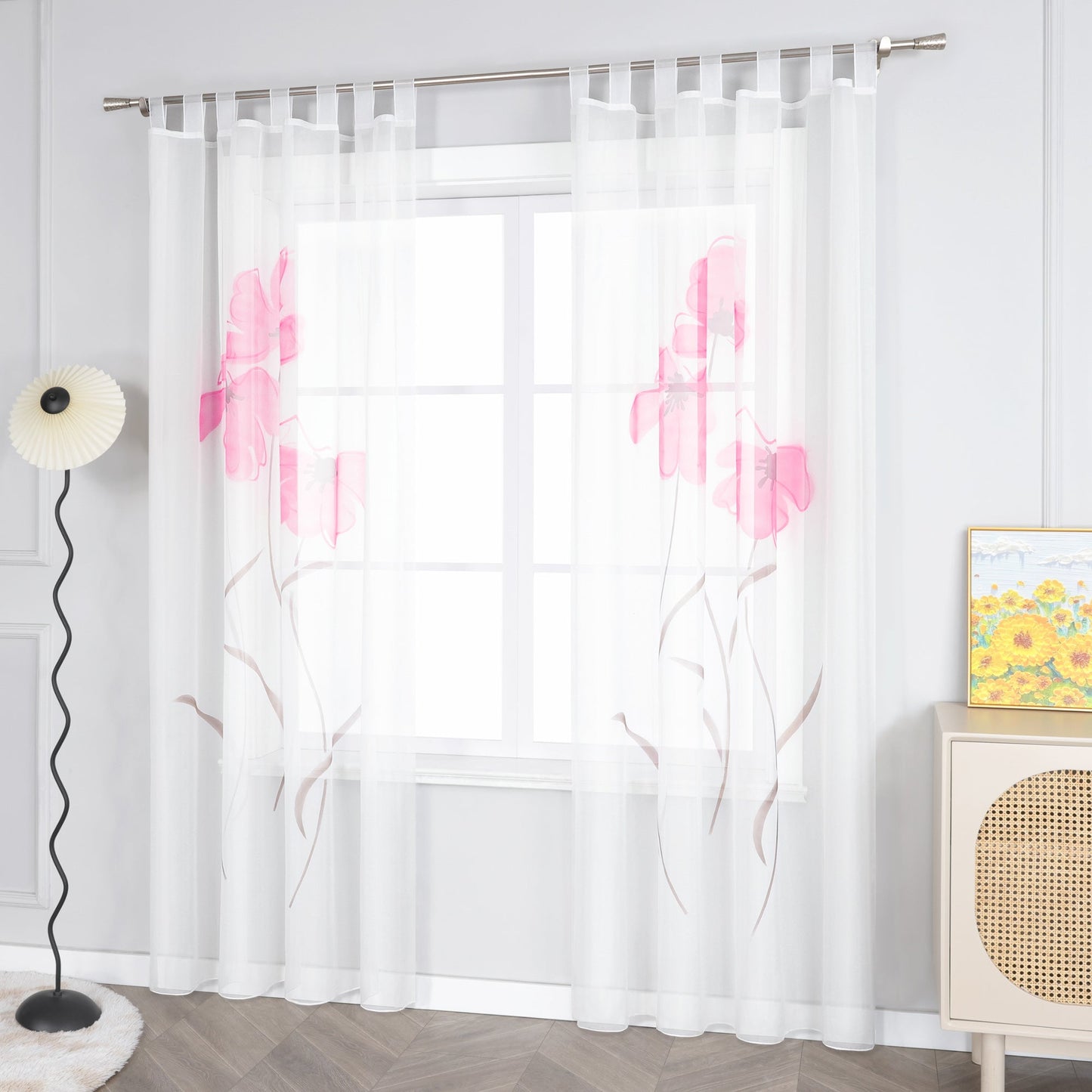 Das Bild zeigt einen weißen Voile-Vorhang mit einem Muster aus rosa Blumen und braunen Zweigen, der mit Metallringen an einer Stange aufgehängt ist. Neben dem Fenster steht eine Stehlampe mit einem weißen Lampenschirm. Daneben befindet sich ein heller Schrank mit einem gewobenen Musterelement auf der Oberfläche, auf dem ein Gemälde in Gelbtönen platziert ist.