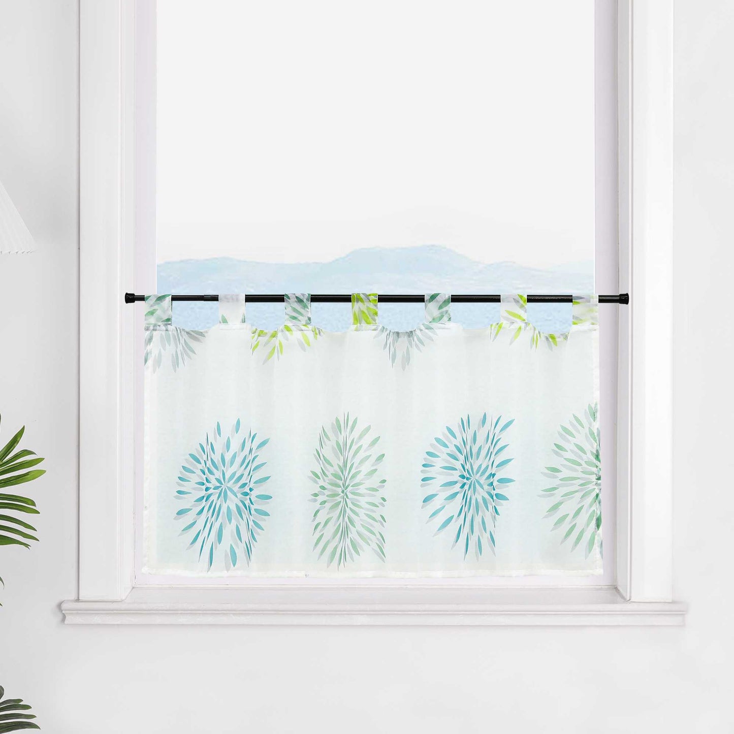 Die transparente Scheibengardine mit grünen und blauen Feuerwerksmotiven hängt dekorativ vor einem Fenster. Die Wand im Hintergrund ist in weiß gehalten, wodurch die bunten Feuerwerksmuster auf dem Stoff besonders zur Geltung kommen. Die Gardine verleiht dem Raum eine festliche, fröhliche Atmosphäre.