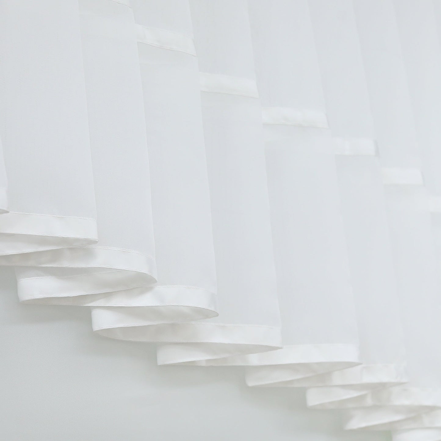 Dies ist ein Detailbild eines weißen Vorhangs, der aus leichtem, transparentem Stoff besteht. Der Vorhang ist in wellenförmigen, mehrschichtigen Lagen angeordnet, mit sorgfältig verarbeitetem Saum, der ihm Tiefe und dekorative Wirkung verleiht. Ideal für die Fensterdekoration