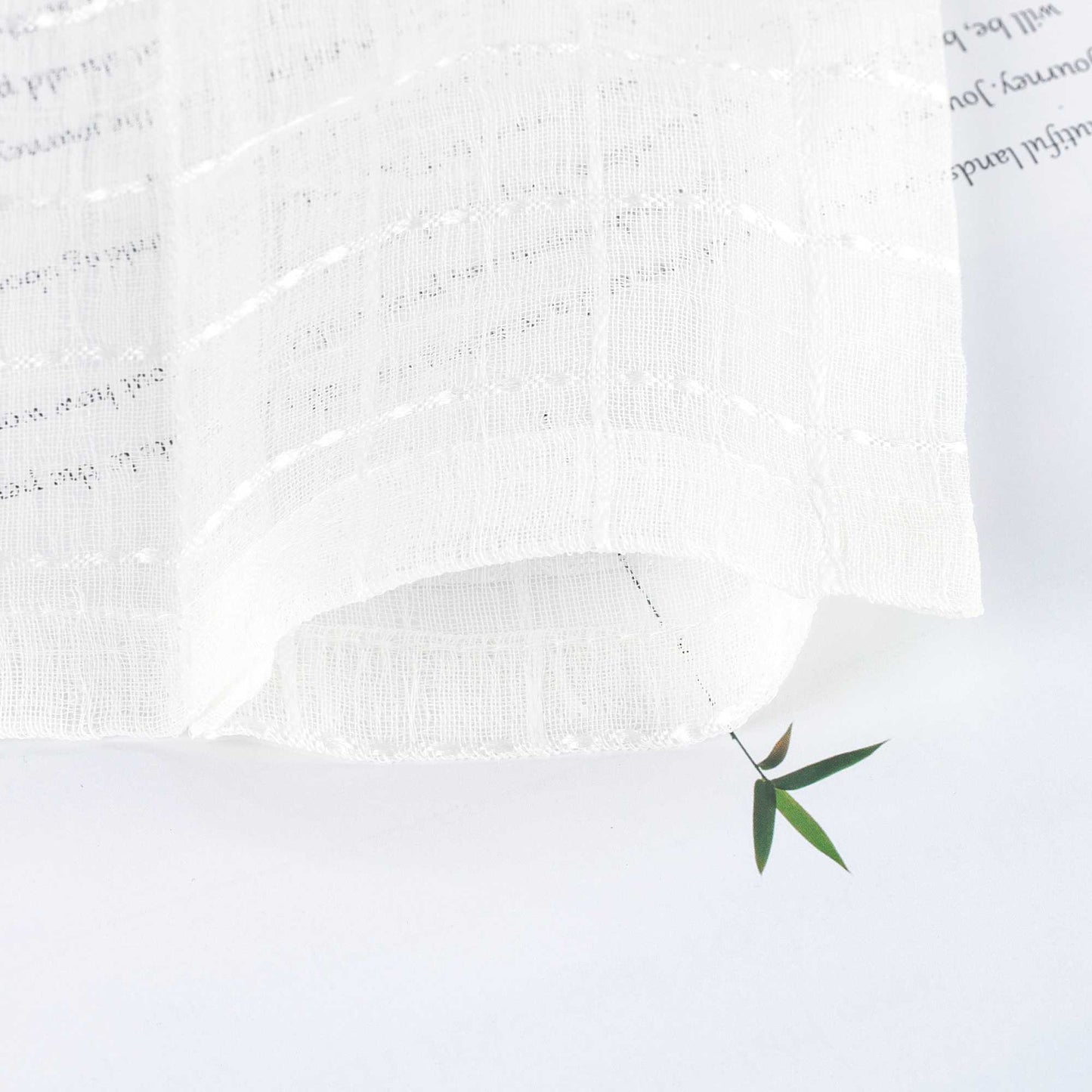 Nahaufnahme eines weißen, transparenten Vorhangstoffs mit feiner Gitterstruktur. Der Stoff fällt weich und wirkt luftig-leicht. Ein grünes Blatt darunter betont die Lichtdurchlässigkeit.