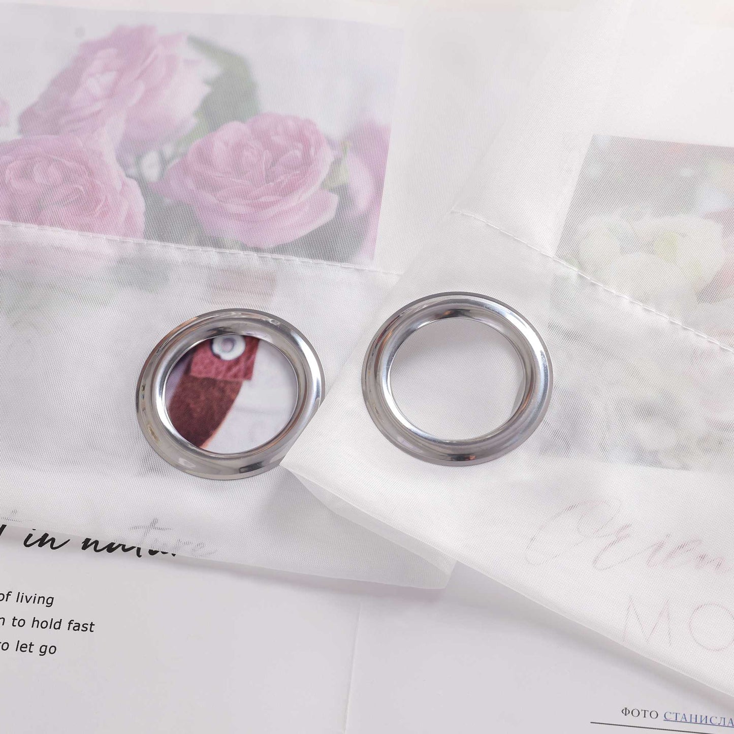 Der weiße Vorhangstoff auf dem Bild ist mit zwei Metalllöchern versehen. Durch den hellen und transparenten Stoff ist ein florales Muster im Hintergrund schwach zu erkennen. So wie es aussieht, sind die Ösen wahrscheinlich aus rostfreiem Stahl gefertigt.