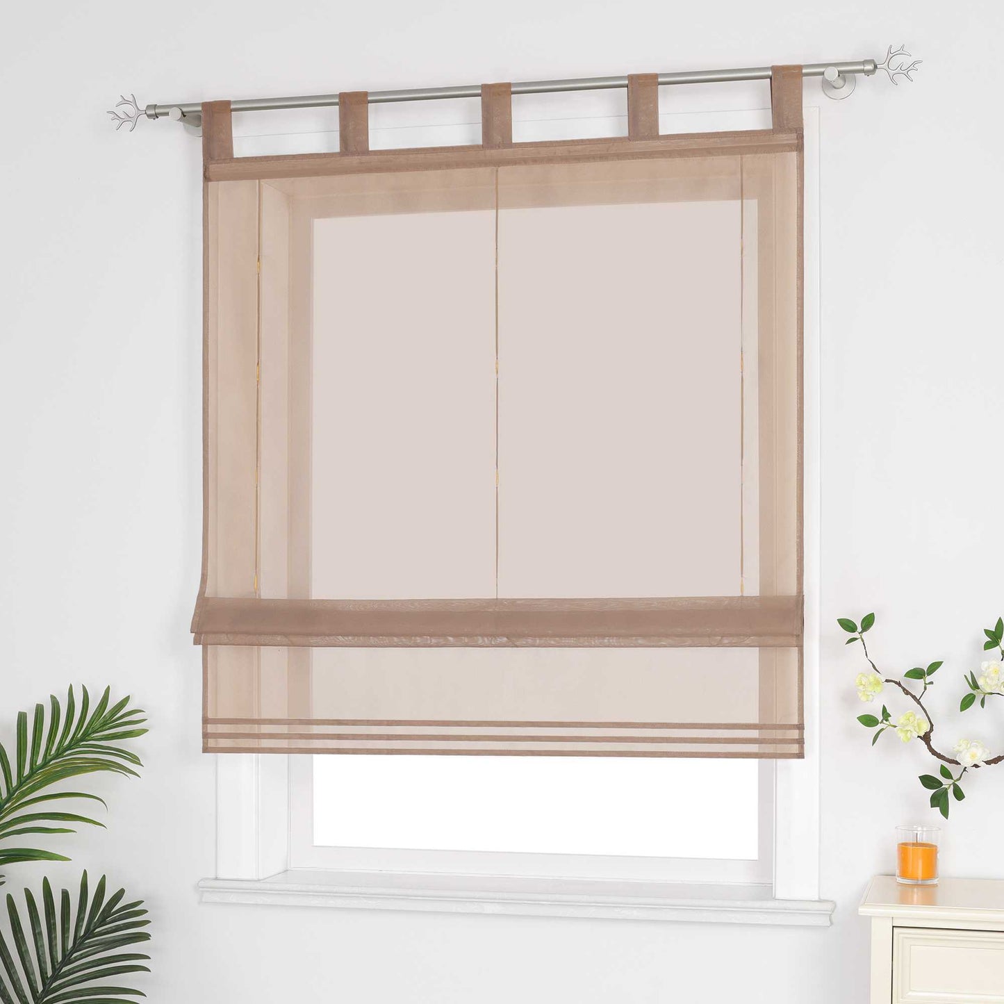 Halbtransparenter brauner Raffrollo hängt vor dem Fenster und sorgt für eine helle, luftige Atmosphäre. Daneben setzen grüne Pflanzen dekorative Akzente. Seitliche Ansicht.