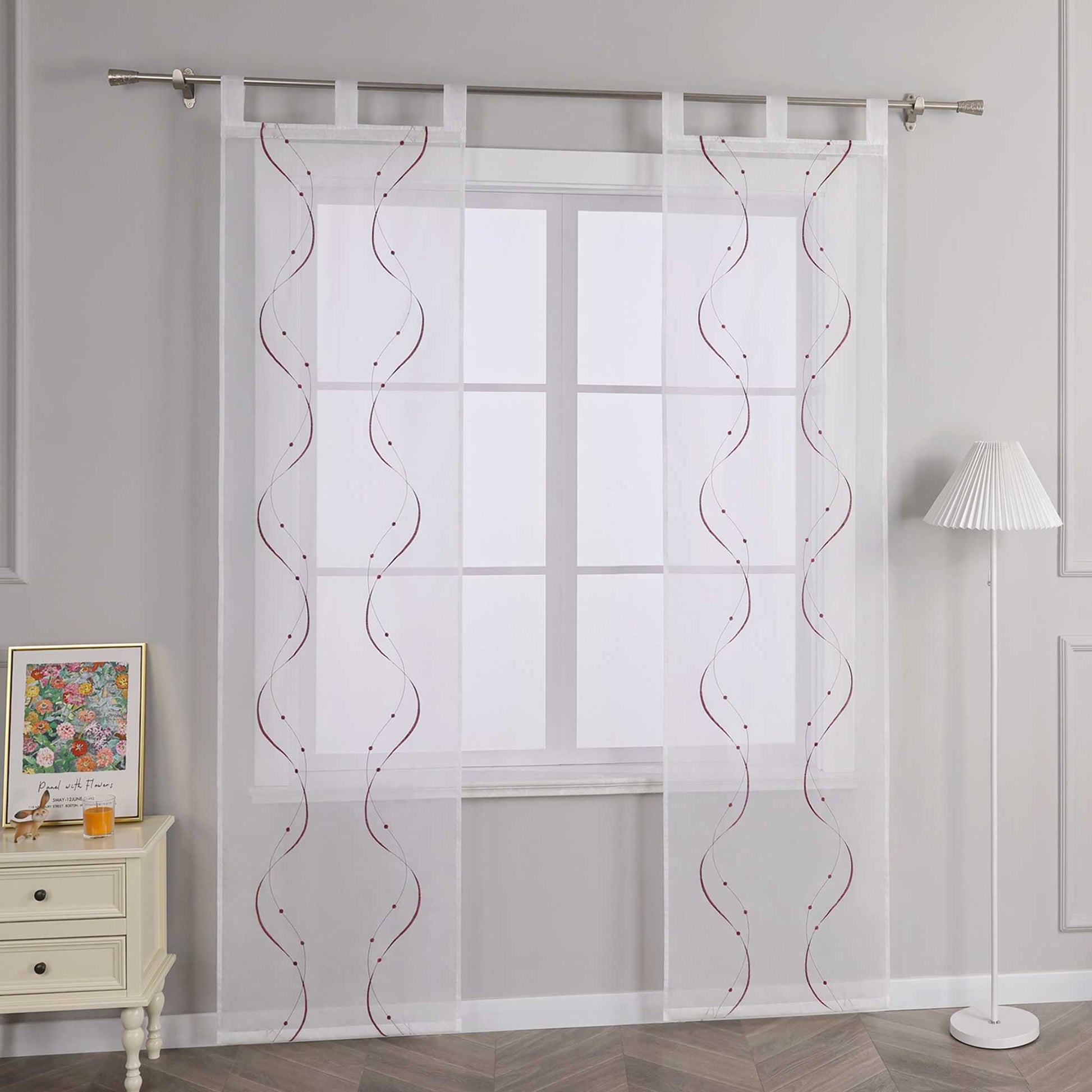 Joyswahl – Das Bild zeigt die Seitenansicht einer weissen transparenten Gardine mit Schlaufen, die an einer Metallstange haengt. Die Gardine ist mit lila Wellenperlenmuster und Stickerei versehen. Links vom Fenster steht ein heller Schrank mit Schubladen, auf dem ein farbenfrohes Blumenbild und ein orangefarbenes Objekt platziert sind. Rechts daneben steht eine Stehlampe mit weiss gestreiftem Lampenschirm.