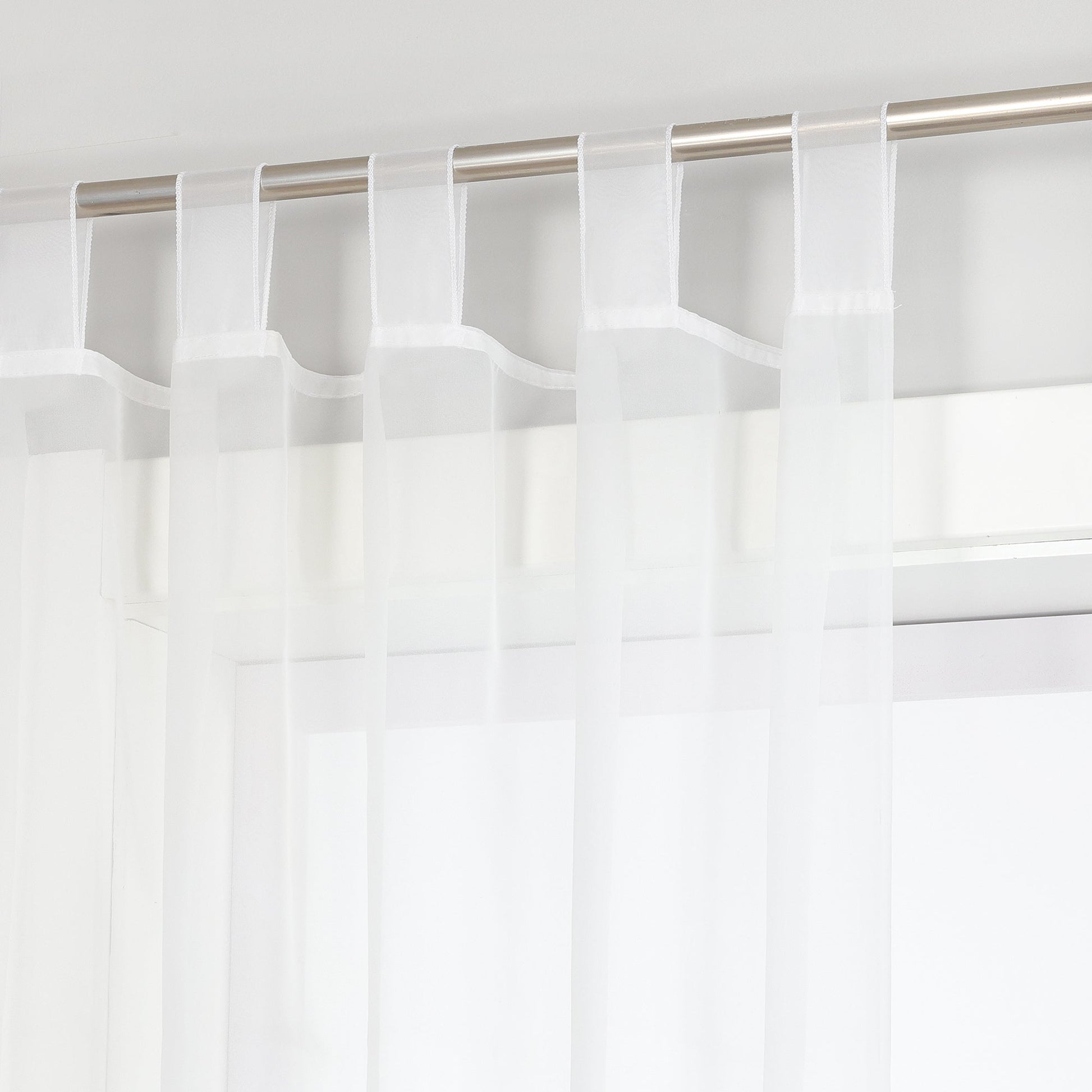 Das Bild zeigt eine Vorhangstange, an der weiße Voile-Vorhänge hängen. Die Vorhänge sind mit Haken an der Stange befestigt, und durch die Voile-Vorhänge ist ein Teil des Fensters sichtbar. Insgesamt vermittelt das Bild einen einfachen und hellen Eindruck.