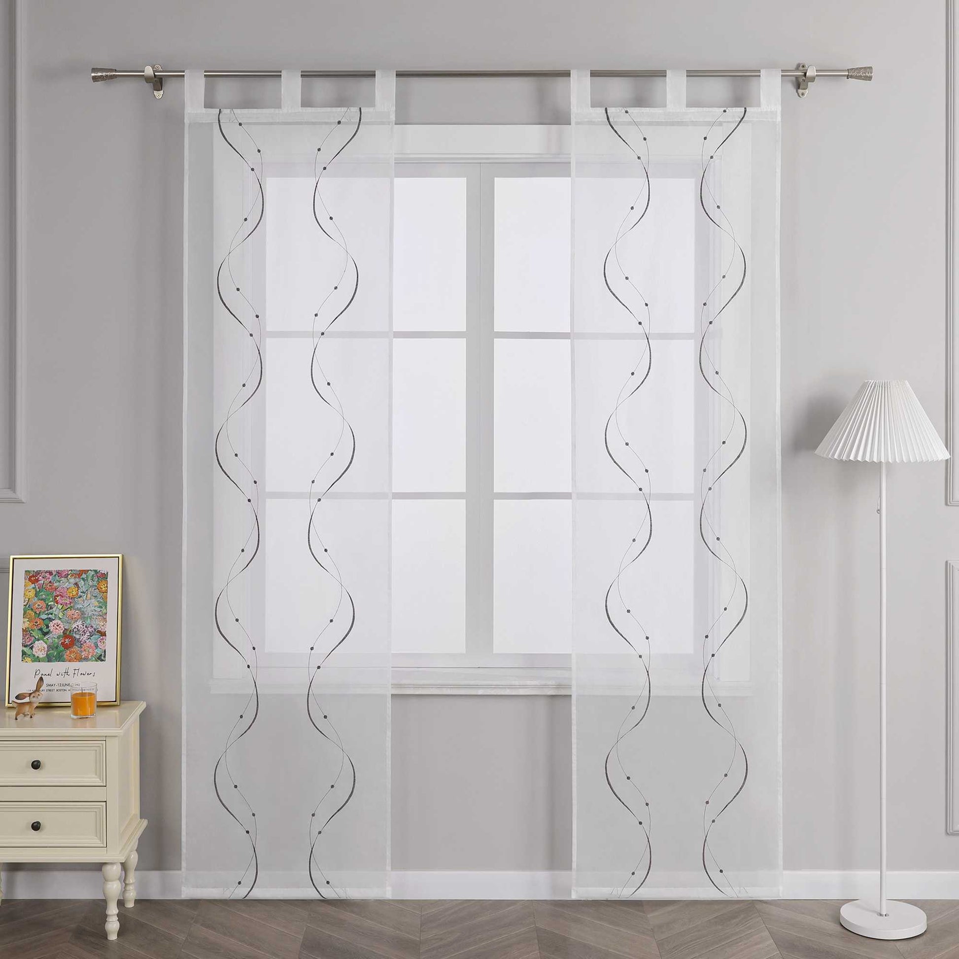Vor einer hellgrauen Wand befindet sich ein Fenster, an dem eine weisse transparente Gardine mit grauem symmetrischem Wellenperlenmuster und Stickerei haengt. Die Gardine ist an einer Metallstange mit Schlaufen befestigt. Links vom Fenster steht ein heller Schrank mit Schubladen, darauf ein farbiges Blumenbild und ein kleines orangefarbenes Objekt. Rechts vom Fenster steht eine Stehlampe mit weissem gestreiftem Lampenschirm.