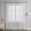 Vor einer hellgrauen Wand befindet sich ein Fenster, an dem eine weisse transparente Gardine mit grauem symmetrischem Wellenperlenmuster und Stickerei haengt. Die Gardine ist an einer Metallstange mit Schlaufen befestigt. Links vom Fenster steht ein heller Schrank mit Schubladen, darauf ein farbiges Blumenbild und ein kleines orangefarbenes Objekt. Rechts vom Fenster steht eine Stehlampe mit weissem gestreiftem Lampenschirm.