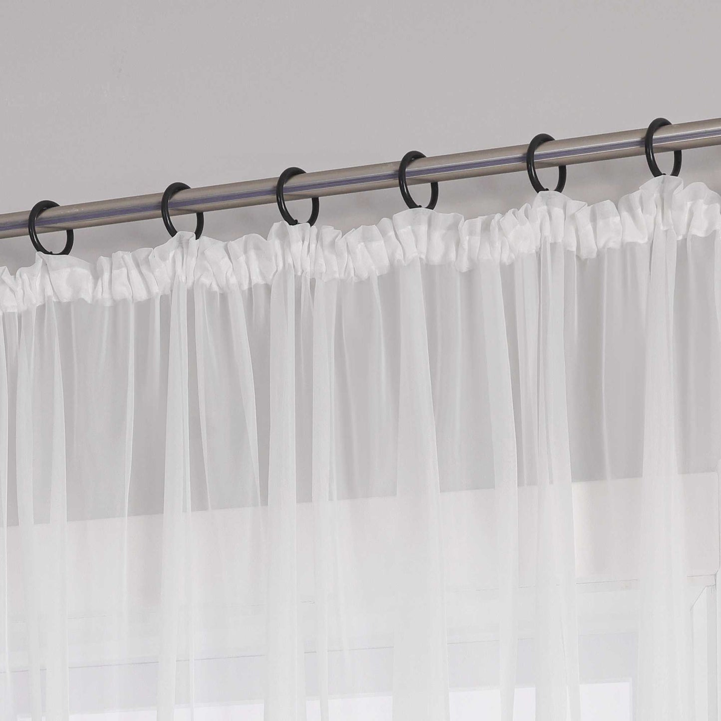 Eine Metallgardinenstange mit schwarzen Ringen, an denen ein weißer Vorhang mit Kräuseln aus transparentem Stoff aufgehängt ist. Der Vorhang hat am oberen Ende ein gerafftes Design.
