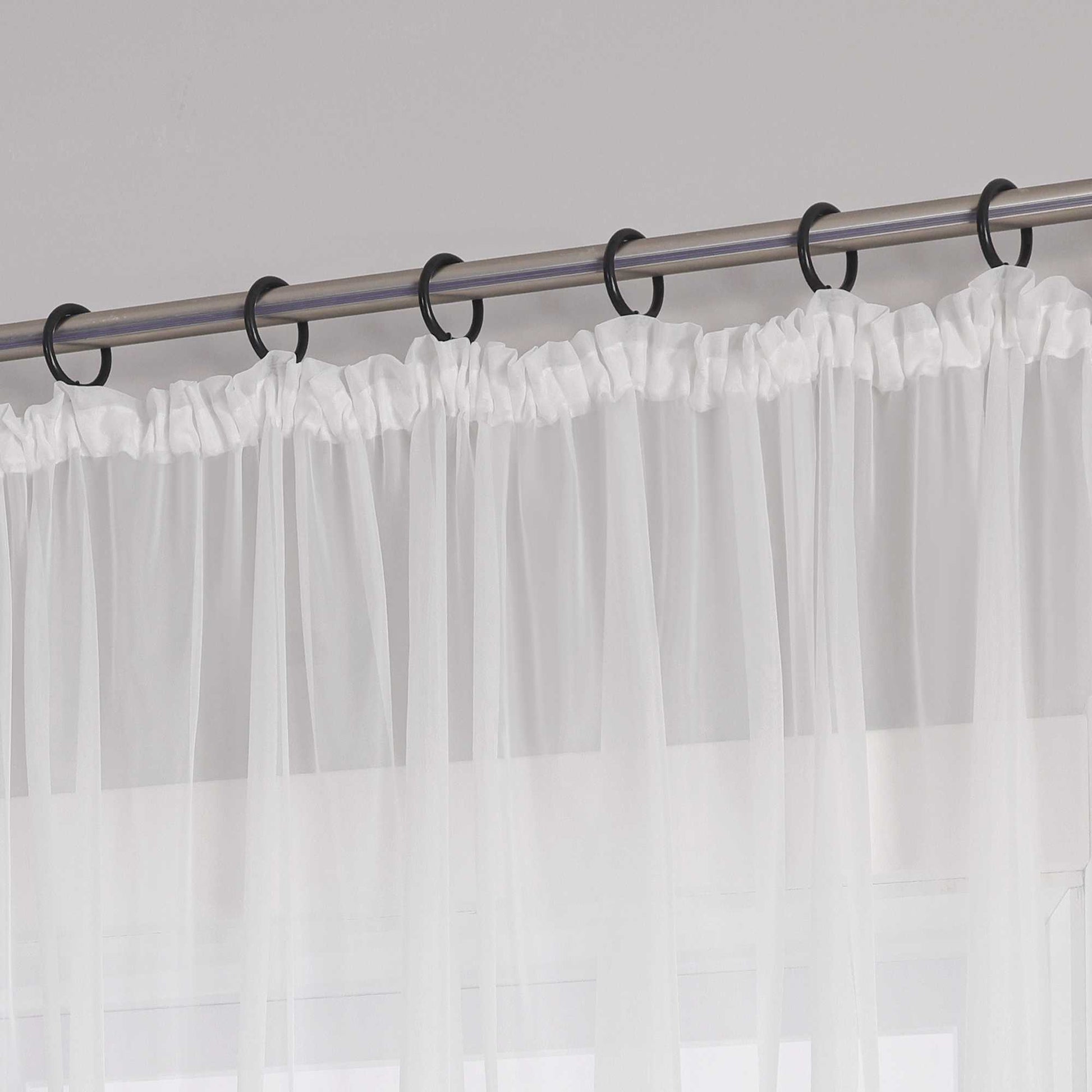 Eine Metallgardinenstange mit schwarzen Ringen, an denen ein weißer Vorhang mit Kräuseln aus transparentem Stoff aufgehängt ist. Der Vorhang hat am oberen Ende ein gerafftes Design.