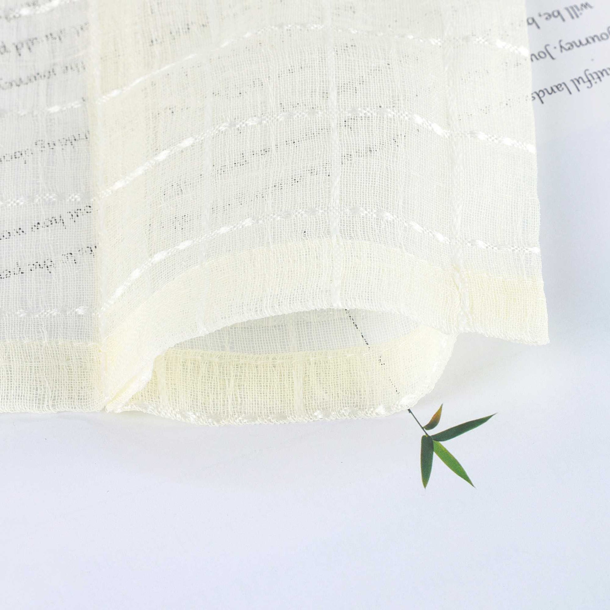 Der cremeweiße Vorhangstoff liegt locker gebogen auf einer hellen Fläche. Die feine Karostruktur und luftige Transparenz sind deutlich erkennbar. Ein grünes Blatt unter dem Stoff betont die weiche Textur und Lichtdurchlässigkeit.