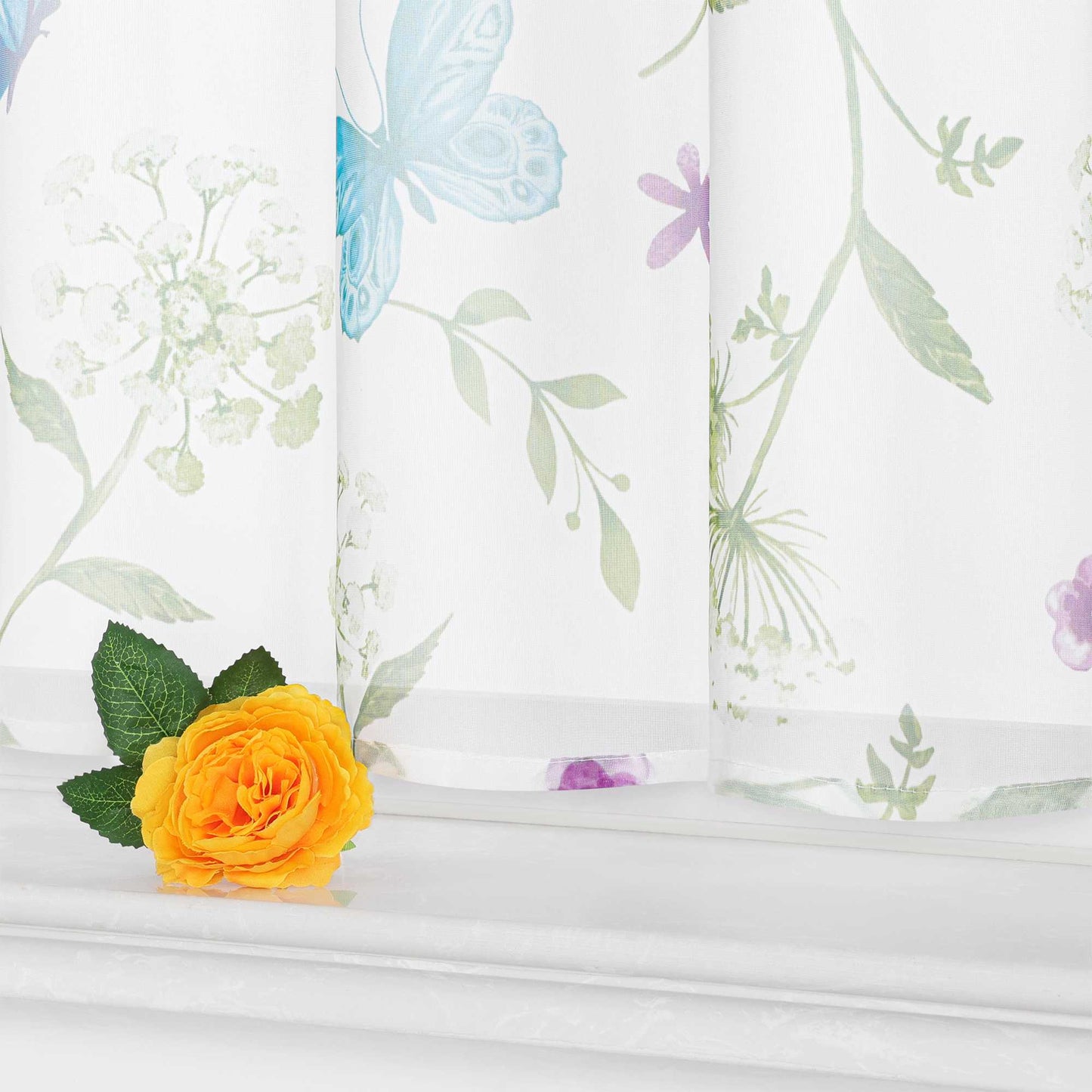 Detail der weißen Scheibengardine am unteren Rand, mit blauen Schmetterlingen, grünen Zweigen, weißen Blumen und zarten lila Akzenten. Auf dem Fensterbrett steht eine gelbe Kunstblume mit grünen Blättern.