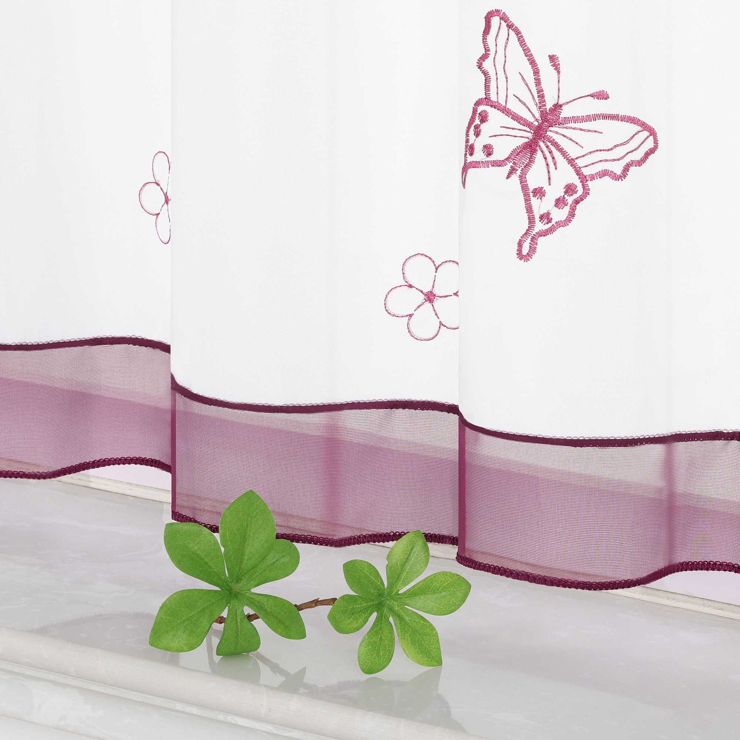 Nahaufnahme des unteren Teils eines lila transparenten Vorhangs mit Schmetterlings- und kleinen Blumenmustern. Der Vorhang ist mit einem lila Dekoband am unteren Rand verziert.