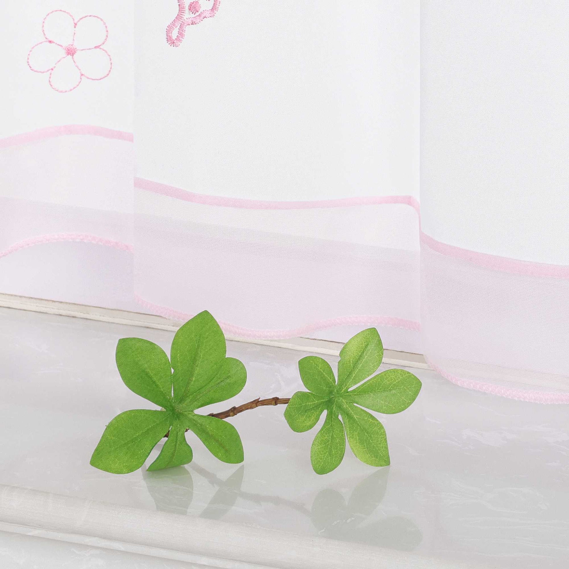 Nahaufnahme des unteren Teils eines rosa transparenten Vorhangs mit Schmetterlings- und kleinen Blumenmustern. Der Vorhang ist mit einem rosa Dekoband am unteren Rand verziert.