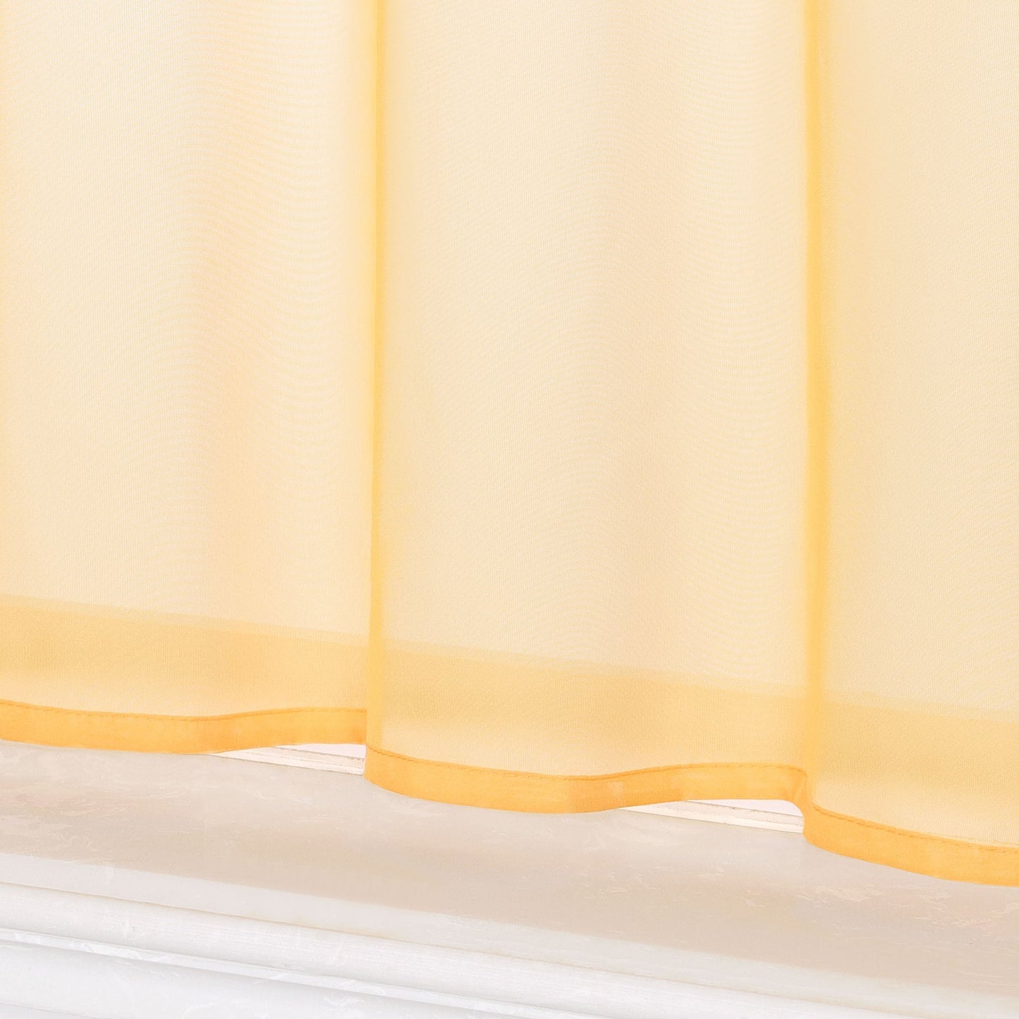 Dies ist eine Nahaufnahme des unteren Teils eines gelben, transparenten Vorhangs. Der Stoff ist leicht und durchsichtig, mit gelbem Saum am unteren Rand und einem weißen Fensterbrett darunter.