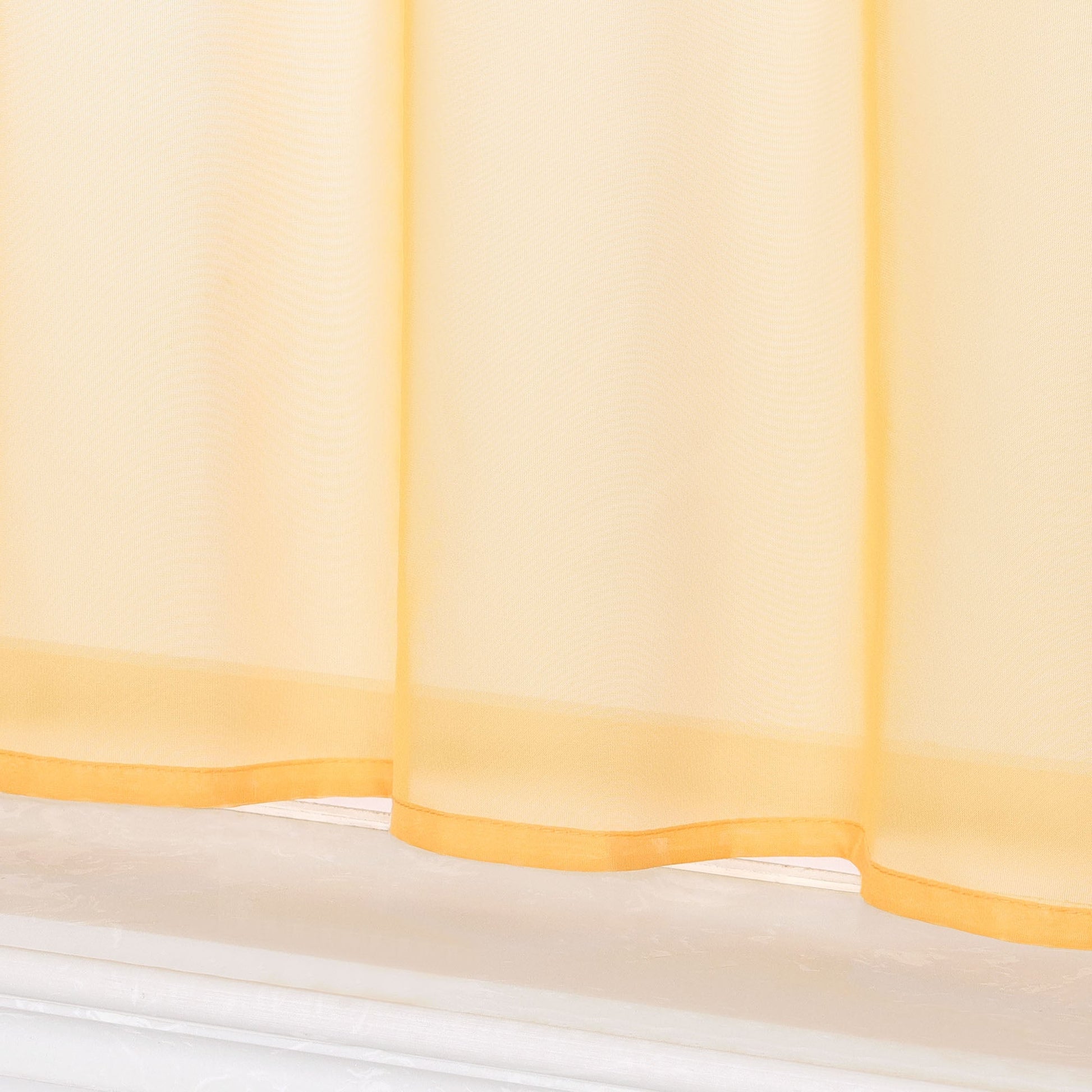 Dies ist eine Nahaufnahme des unteren Teils eines gelben, transparenten Vorhangs. Der Stoff ist leicht und durchsichtig, mit gelbem Saum am unteren Rand und einem weißen Fensterbrett darunter.