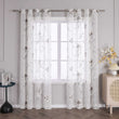 Das Bild zeigt ein Fenster, an dem ein Paar weiße Gardinen hängt. Die Gardinen sind mit einem filigranen Blumenmuster verziert, wobei die Blüten hauptsächlich in Weiß und Braun gehalten sind. Die Blütenmitte ist mit roten Akzenten verziert, und die Zweige sind in schwarzen Linien dargestellt. Die Gardinen sind an einer Metallstange befestigt, die an beiden Enden an einer hellgrauen Wand montiert ist.