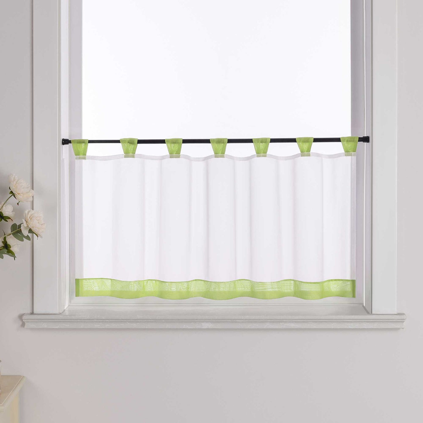 Die gruene Scheibengardine hängt vor einem Fenster, wobei die Hintergrundwand weiß ist. Der Vorhang ist transparent und wird von blumigen Dekorationen an der Seite begleitet.