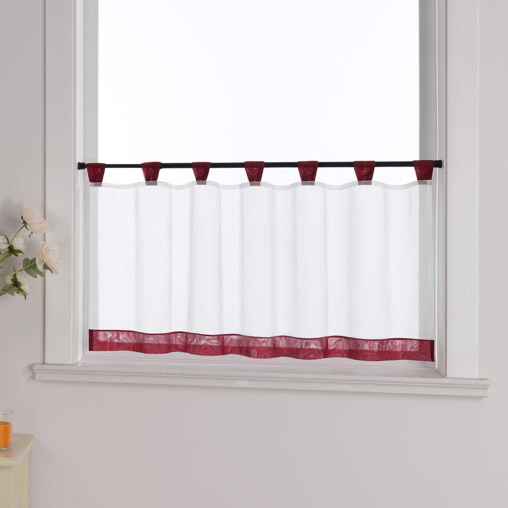 Die weinrot Scheibengardine hängt vor einem Fenster, während die weiße Hintergrundwand im Hintergrund sichtbar bleibt. Der transparente Vorhang lässt das Licht durch, während er von blumigen Dekorationen an der Seite sanft ergänzt wird, was dem Raum eine leichte und frische Atmosphäre verleiht.